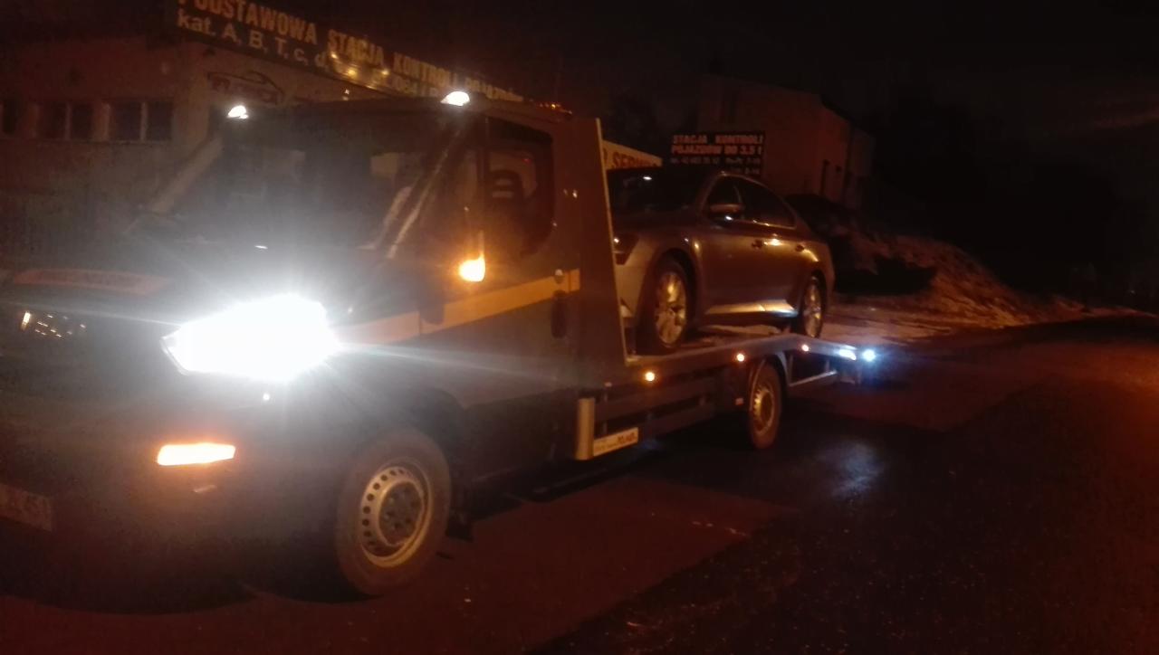 Laweta z szarym samochodem osobowym na platformie, sfotografowana nocą na tle budynku Stacji Kontroli Pojazdów, reflektory lawety oślepiają.