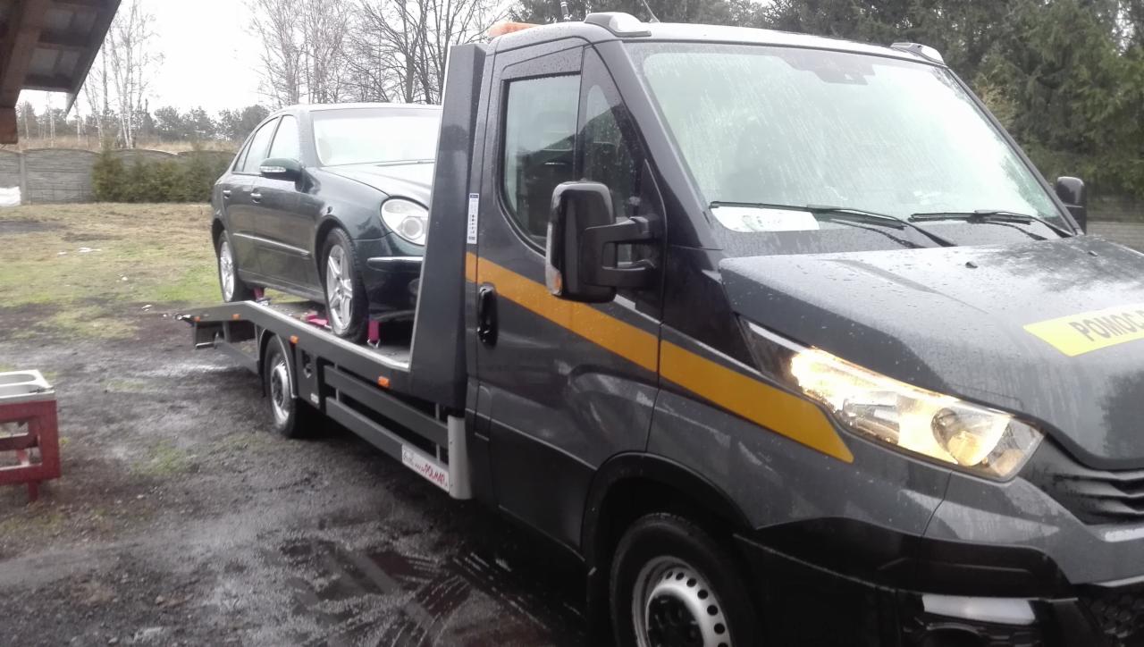 Ciemnoszary samochód osobowy marki Mercedes na platformie lawety z żółtymi pasami, widok z boku na zewnątrz w pochmurny dzień.