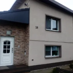 Dwukondygnacyjny budynek z beżową elewacją, fragmentami wykończonymi kamieniem dekoracyjnym, białymi oknami z brązowymi obramowaniami i numerem domu '18' nad oknem. Wejście z czarnym zadaszeniem...