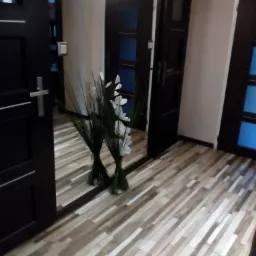 Korytarz z podłogą z paneli imitujących drewno, widoczne trzy ciemne drzwi z przeszkleniami i dekoracyjne bukiety kwiat&oacute;w odbijające się w lustrze.