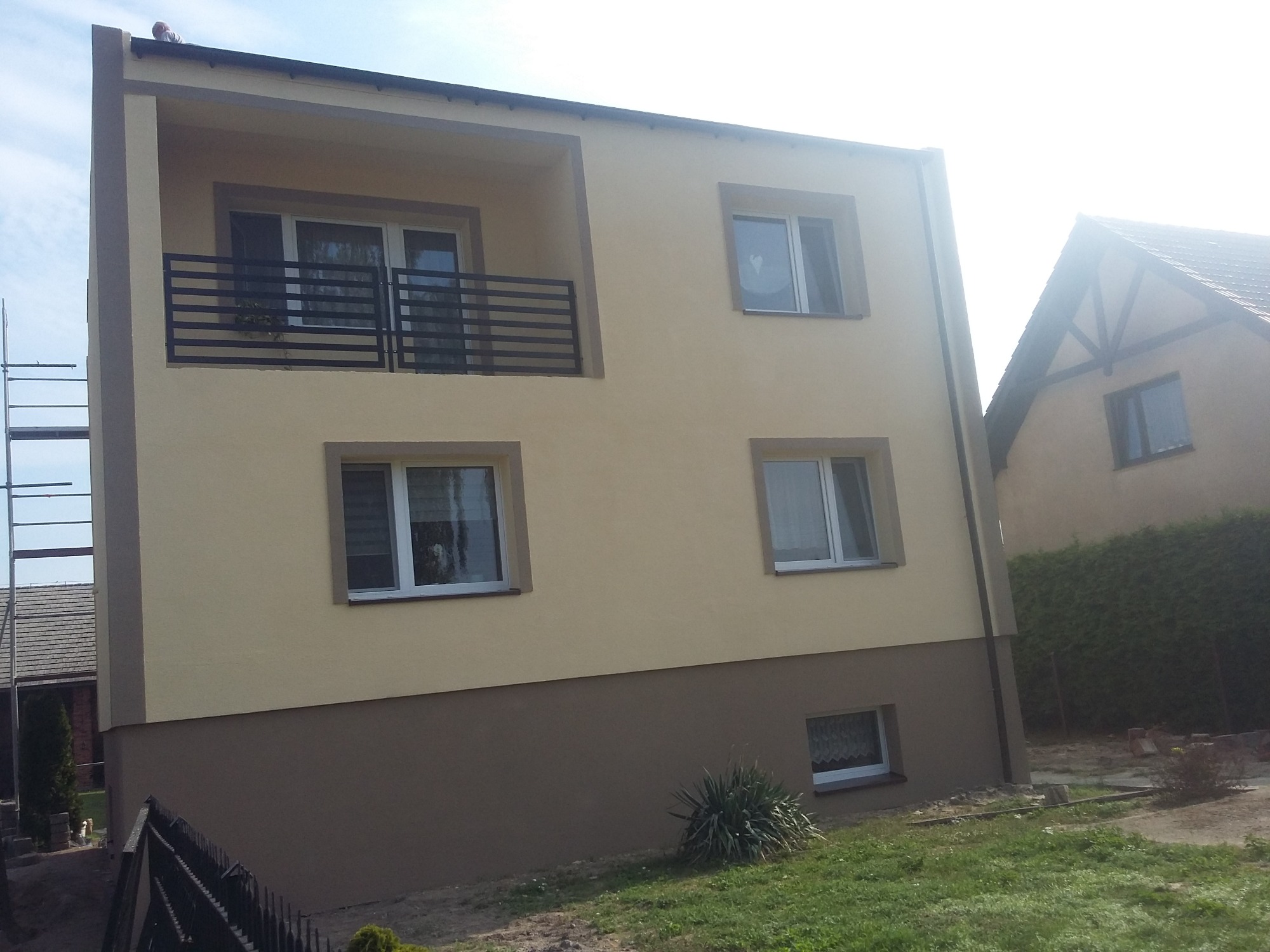 Dwukondygnacyjny dom z beżowo-brązową elewacją, balkonem z czarną balustradą i białymi oknami, widoczny w słoneczny dzień z perspektywy niskiego kąta.