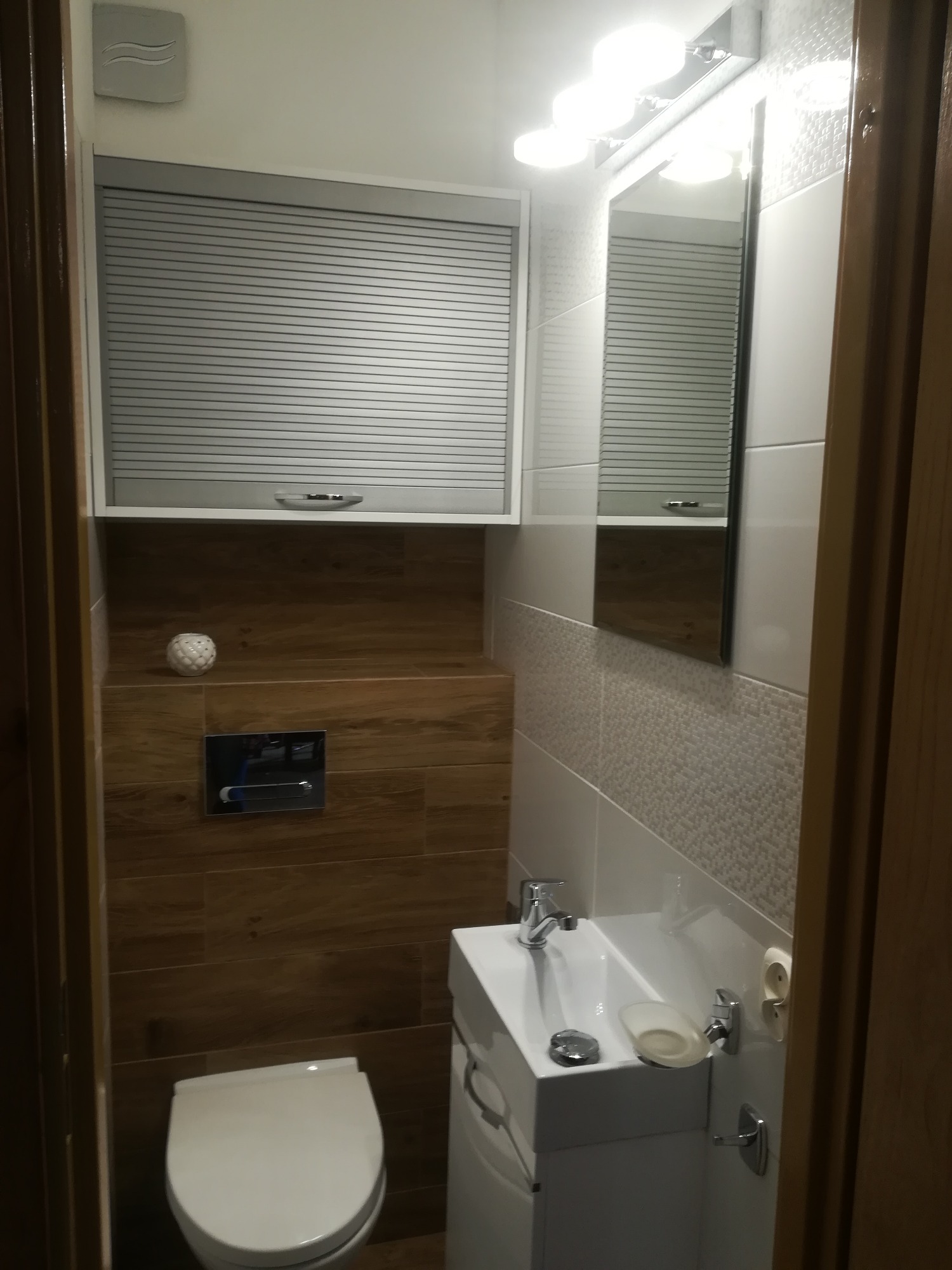 Nowoczesna toaleta z drewnopodobnymi płytkami, szafką z roletą i minimalistyczną umywalką.