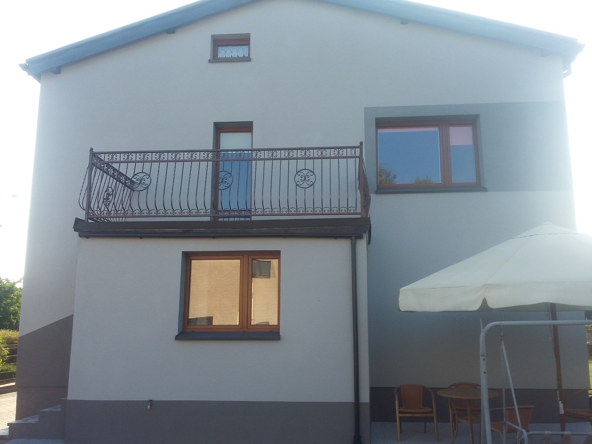Dwupiętrowy dom z jasnoszarą elewacją, ciemnoszarą dolną częścią i balkonem z ozdobną, metalową balustradą, okna z drewnianymi ramami, widoczny parasol ogrodowy i meble.
