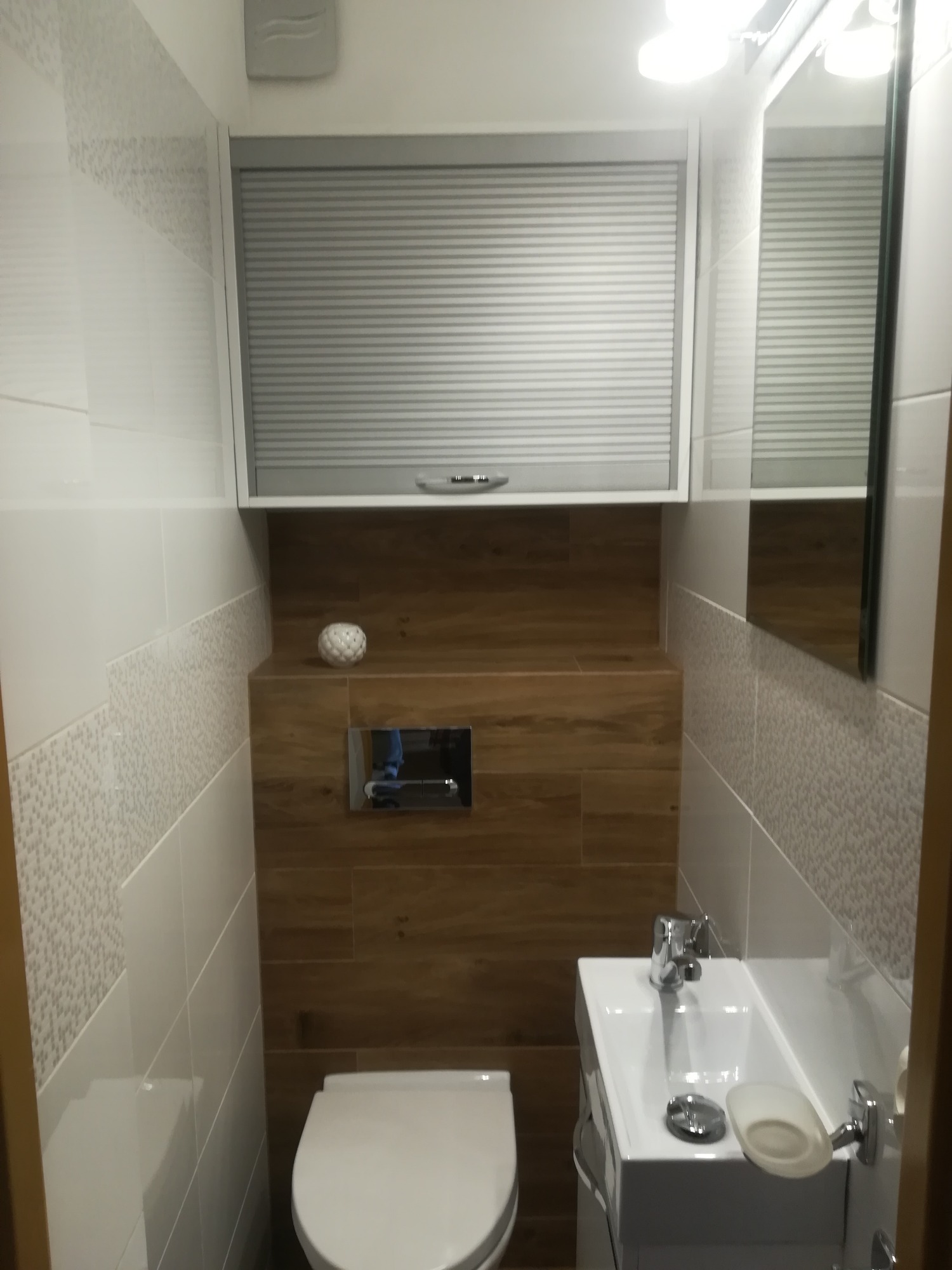 Nowoczesna, biało-brązowa łazienka z podwieszaną toaletą, umywalką nablatową i szafką z roletą, wykończona płytkami imitującymi drewno i dekoracyjnymi płytkami ściennymi.