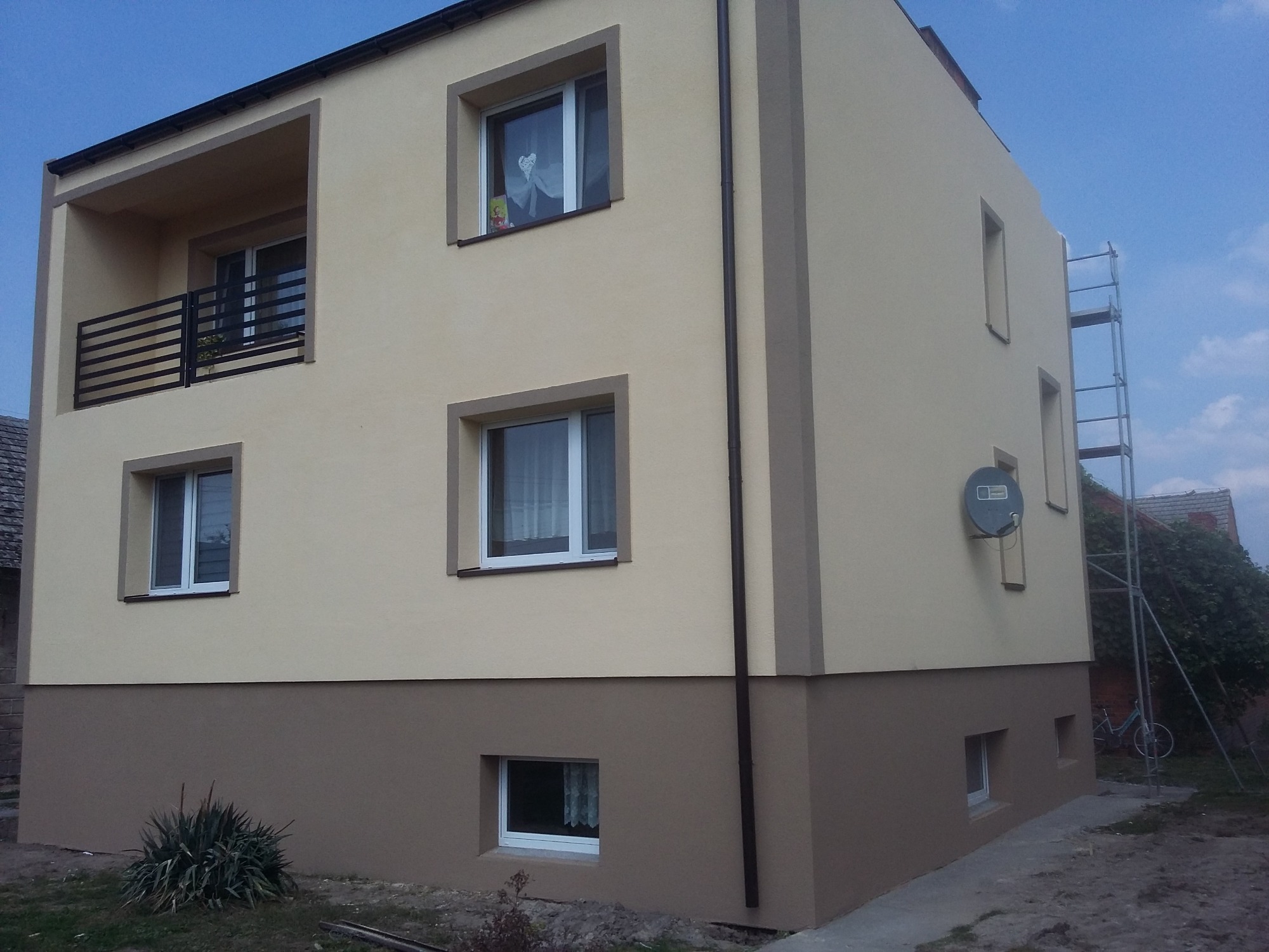 Dwukondygnacyjny dom z balkonem, otynkowany i pomalowany na beżowo z brązowym cokołem, widoczne rusztowanie przy bocznej ścianie, antena satelitarna na elewacji.