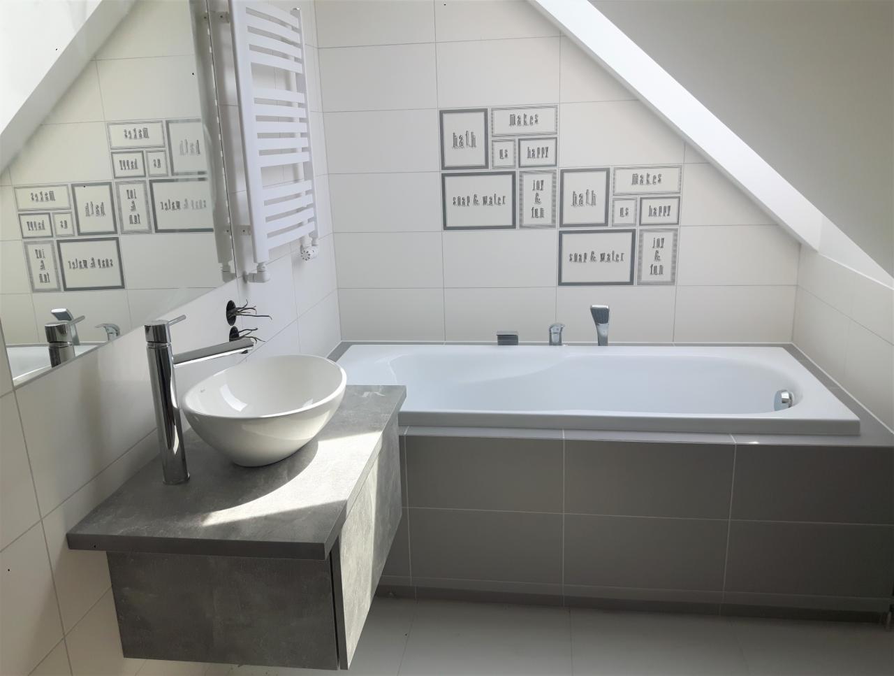 Nowoczesna łazienka na poddaszu ze skośnym sufitem, biała wanna obudowana szarymi płytkami, umywalka nablatowa na szarej szafce, białe płytki z szarymi napisami na ścianie, grzejnik drabinkowy.