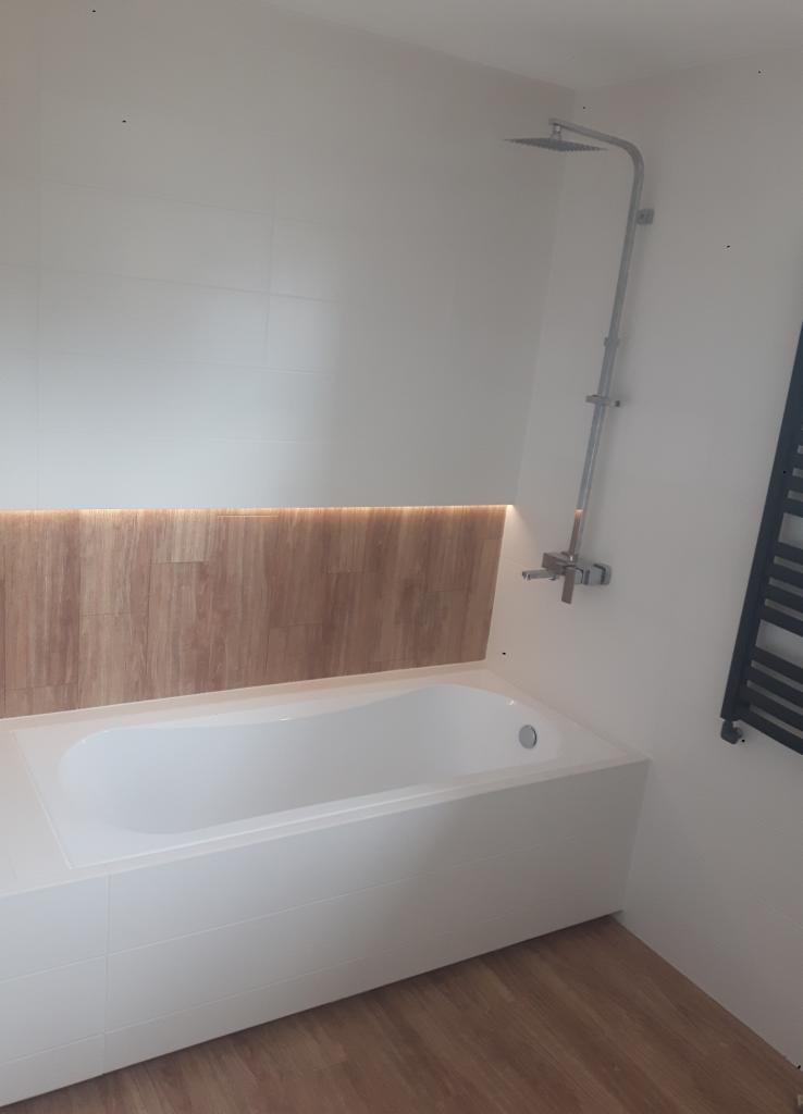 Nowoczesna łazienka z białą wanną obudowaną płytkami, drewnopodobną ścianą z podświetleniem LED oraz chromowaną baterią natryskową.