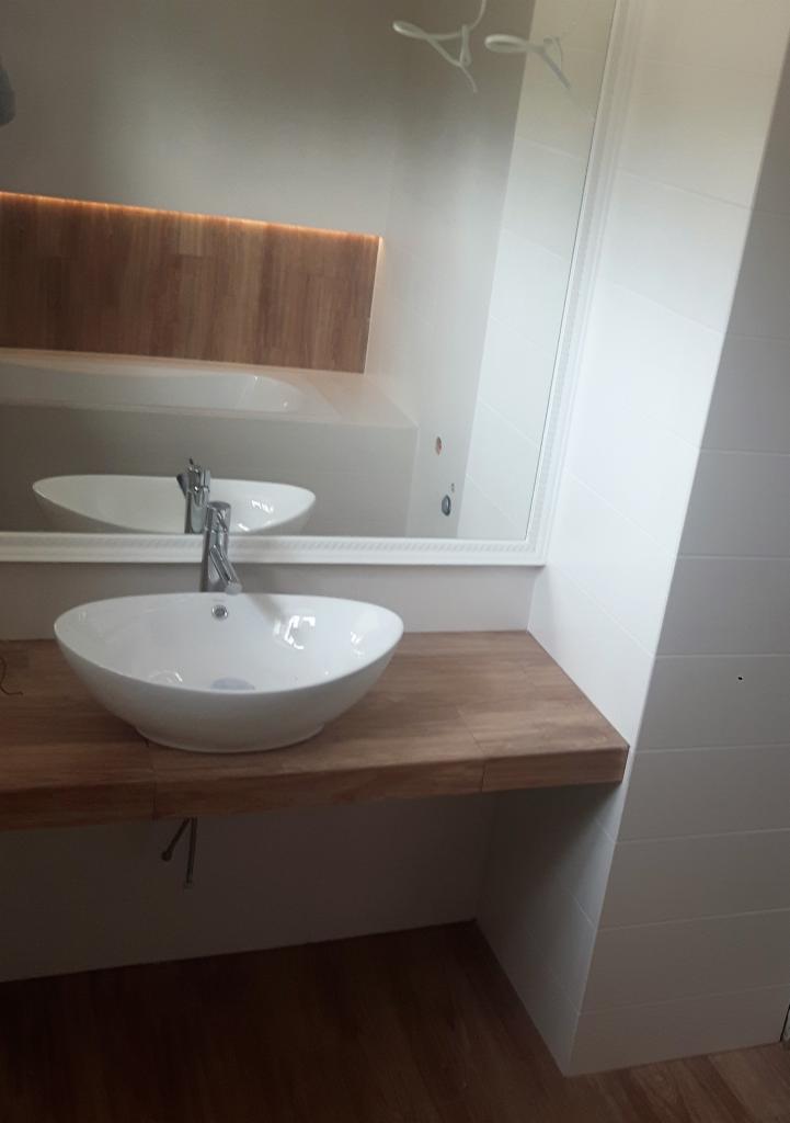 Biała, owalna umywalka nablatowa z chromowaną baterią, osadzona na drewnianym blacie w nowoczesnej łazience z białymi, pionowymi płytkami i dużym lustrem.