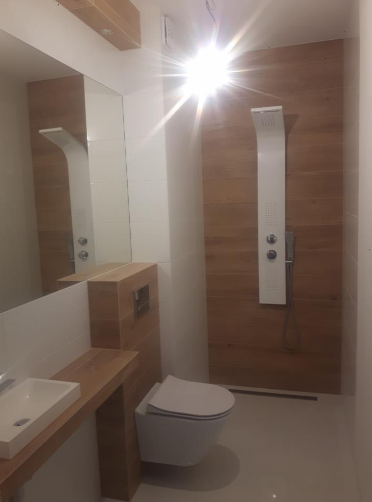 Nowoczesna łazienka z drewnianymi akcentami: panel prysznicowy z hydromasażem na tle ściany wyłożonej płytkami imitującymi drewno, umywalka nablatowa na drewnianym blacie, podwieszana toaleta.