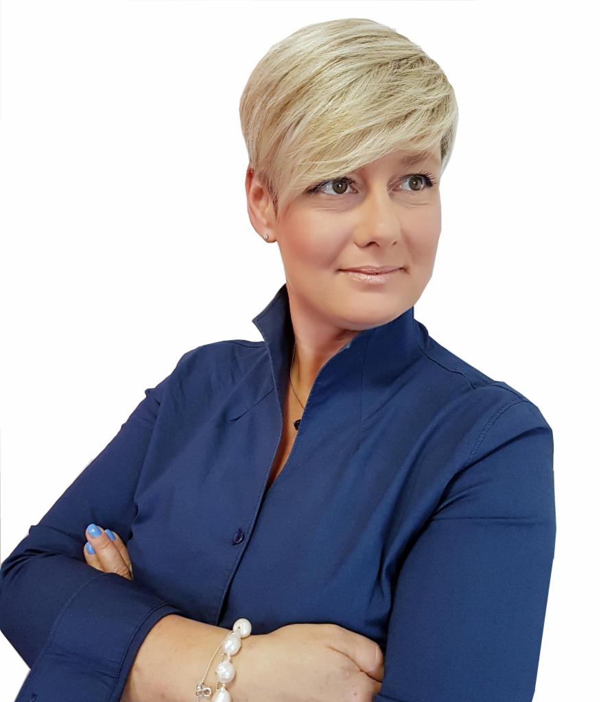 Portret kobiety z krótkimi blond włosami w granatowej koszuli, z założonymi rękami i perłową bransoletką, spoglądającej w bok.