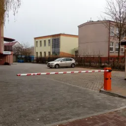 Szary samochód osobowy za szlabanem na brukowanym parkingu przed budynkami, w tle budynek z logo firmy.