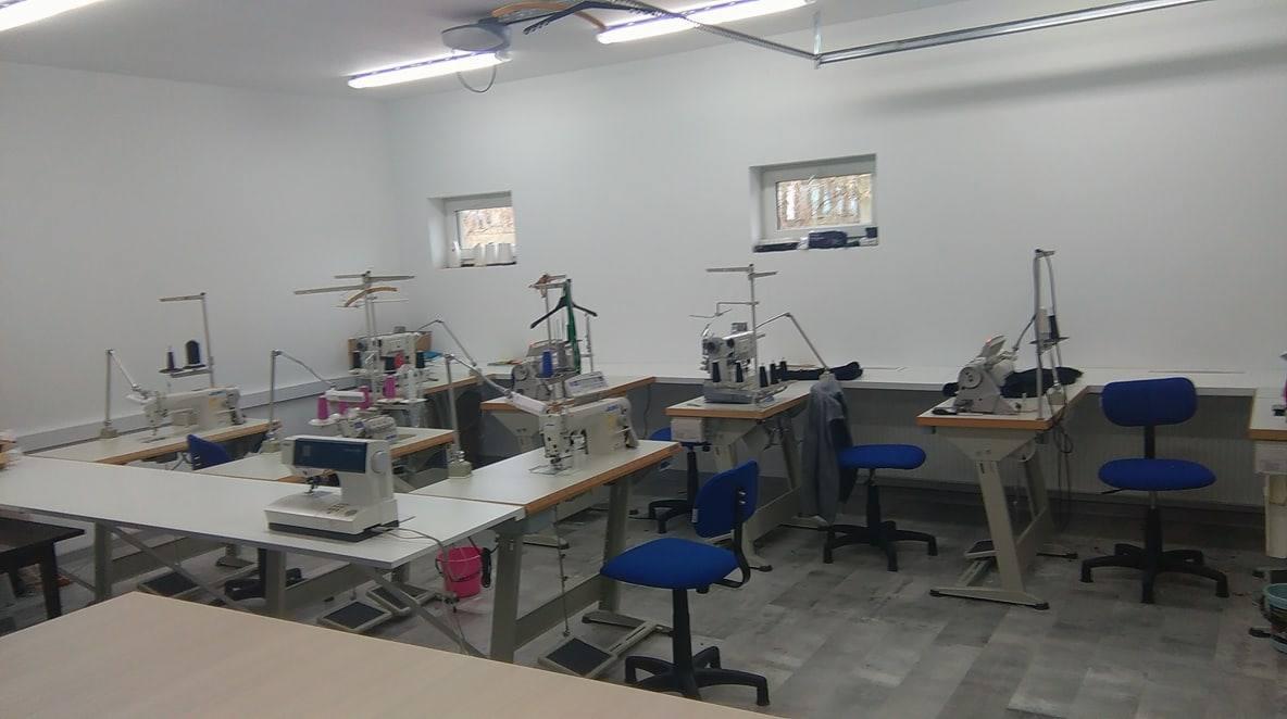 Jasne wnętrze pracowni krawieckiej z rzędem stanowisk wyposażonych w maszyny do szycia, krzesła i akcesoria, oświetlone lampami sufitowymi.