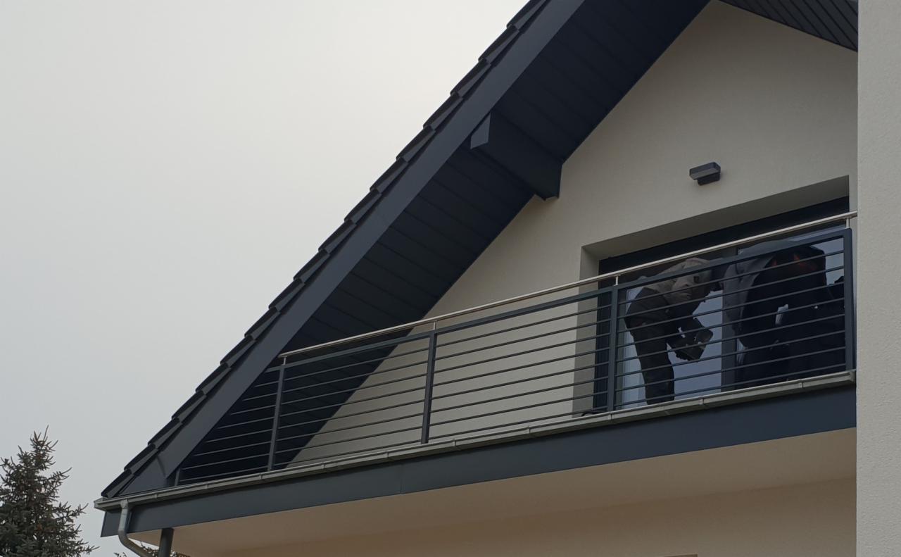 Nowoczesny balkon z poziomą, stalową barierką na tle szarej elewacji domu z ciemną dachówką.