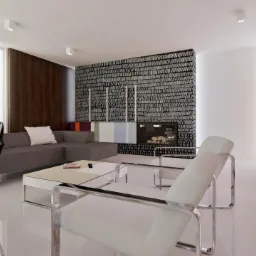 Jasne, przestronne wnętrze salonu z minimalistycznym wystrojem, szarą sofą narożną, białymi fotelami ze stalowym stelażem, ścianą z wzorem imitującym drewno oraz kominkiem obudowanym czarną...