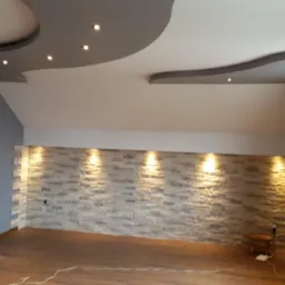 Wykończone wnętrze z ozdobną ścianą z kamienia dekoracyjnego podświetloną punktowymi lampami, podwieszanym sufitem z wbudowanym oświetleniem i drewnopodobną podłogą.