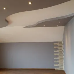 Nowoczesne wnętrze z falistym, dwukolorowym sufitem podwieszanym z oświetleniem punktowym, szarą ścianą i dekoracyjnym obłożeniem kamieniem na ścianach i wokół drzwi.