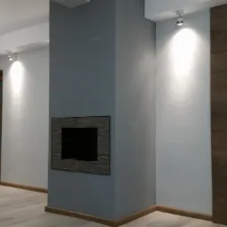 Nowoczesne wnętrze z szarą ścianą, w której wbudowany jest kominek z kamienną obudową, drewniane drzwi z geometrycznymi przeszkleniami i panele ścienne imitujące drewno, oświetlone punktowymi lampami.