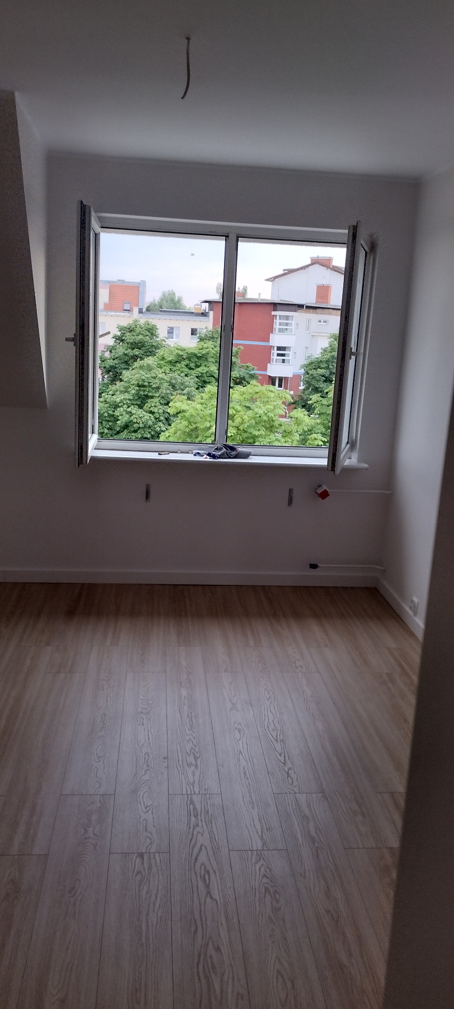 Pusty pokój z drewnianą podłogą i otwartym oknem, widok na zieleń miejską i budynki, ściany świeżo pomalowane na biało, widoczne ślady po demontażu na ścianie pod oknem.