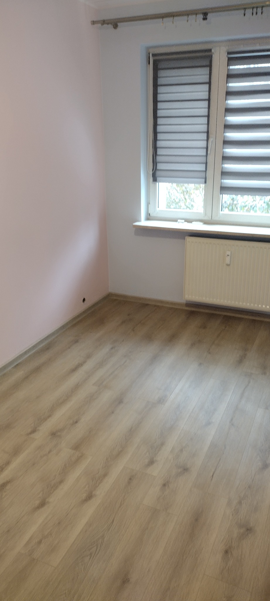 Pusty pokój po remoncie: świeżo pomalowane różowe ściany, jasna podłoga laminowana, białe okno z żaluzjami dzień-noc i grzejnik.