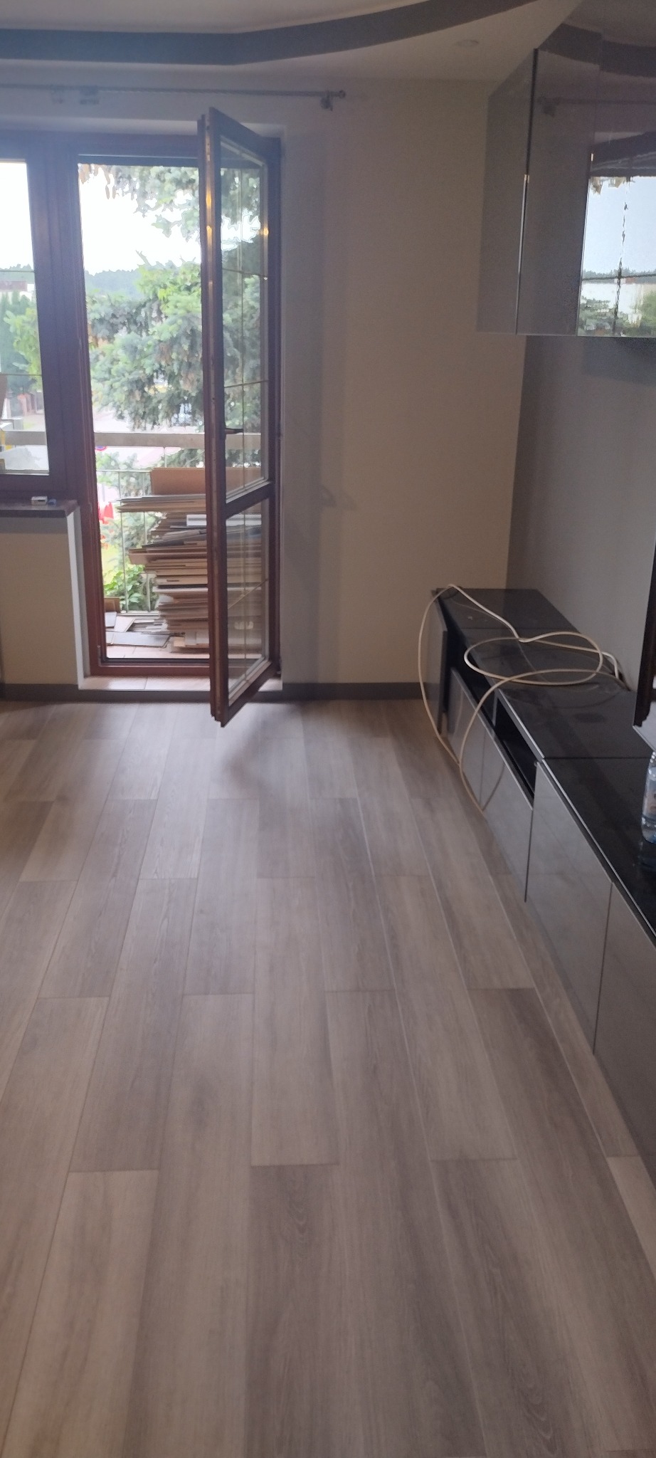 Puste pomieszczenie z szarą podłogą imitującą drewno, otwartym oknem balkonowym wychodzącym na zieleń, oraz niską szafką RTV z czarnym blatem i wystającymi kablami.