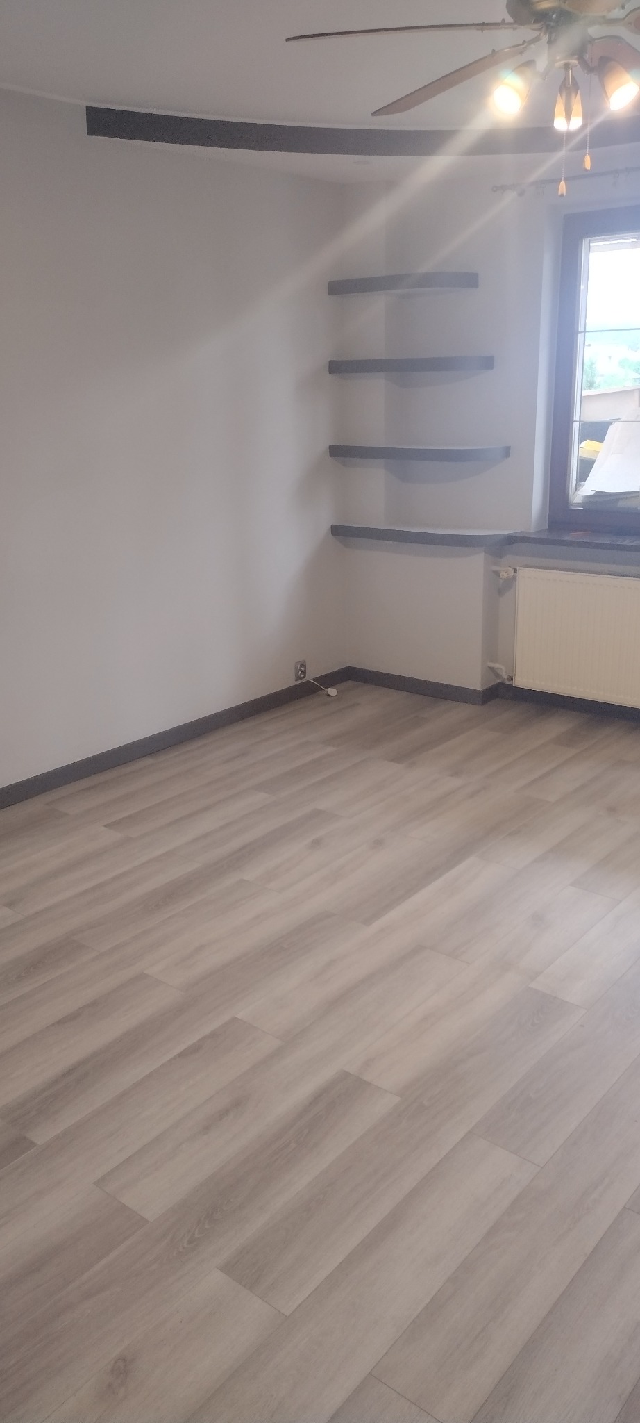 Pomalowane na biało wnętrze pokoju z panelami podłogowymi, trzema półkami w rogu oraz grzejnikiem pod oknem.