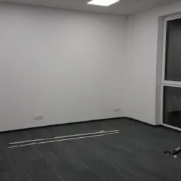 Pomalowane na biało puste biuro z szarą wykładziną, widoczne listwy przypodłogowe i gniazdka elektryczne, oświetlenie sufitowe rastrowe.