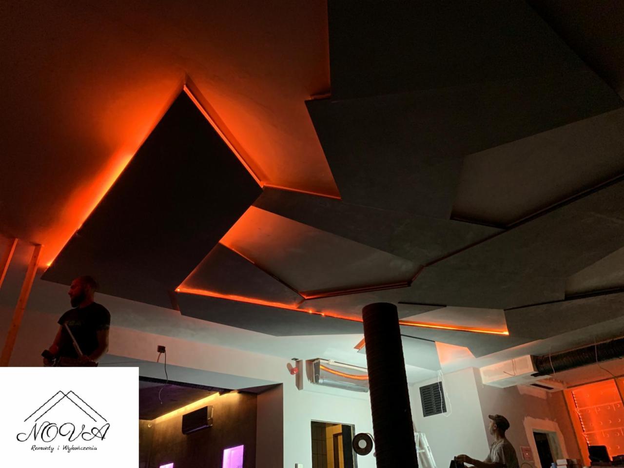 Nowoczesny sufit podwieszany z geometrycznymi panelami i pomarańczowym oświetleniem LED, widoczni dwaj mężczyźni pracujący przy instalacji.