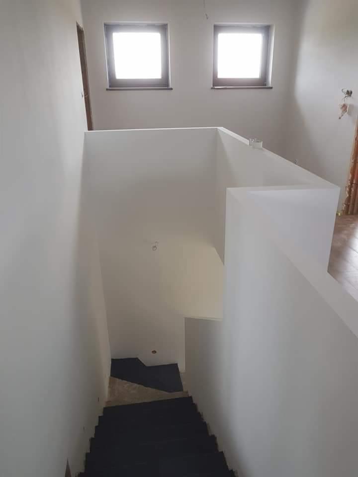 Wnętrze domu w trakcie malowania: białe ściany, schody z ciemnymi stopniami, dwa okna na górze, surowy stan wykończenia.