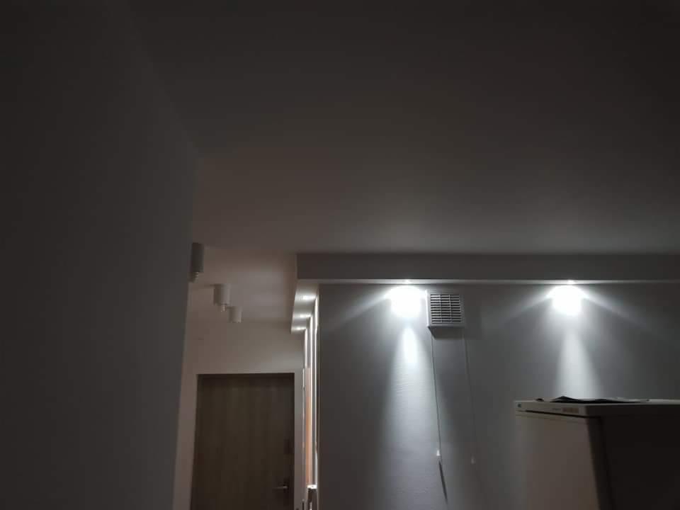 Wnętrze pomieszczenia w trakcie wykańczania, widoczne dwie lampy sufitowe, kratka wentylacyjna, fragment ściany, drzwi i część lodówki, oświetlenie punktowe w suficie, ciemne ujęcie.