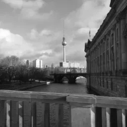 Czarno-białe zdjęcie Berlina z perspektywy mostu, widoczna wieża telewizyjna, rzeka i budynek z kolumnami.
