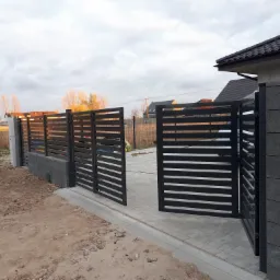 Nowoczesna brama wjazdowa z czarnymi, poziomymi panelami, otwierająca się na wybrukowany podjazd, z ogrodzeniem o podobnym designie i słupkami z szarego betonu architektonicznego w tle krajobraz...