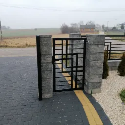 Nowoczesna brama wejściowa z czarnej stali o geometrycznym wzorze, osadzona na szarych, betonowych słupkach. Szara kostka brukowa z żółtym akcentem na podjeździe, w tle widok na pole i zabudowania...