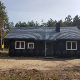 Surowy stan budowy domu szkieletowego z czarną folią izolacyjną, widoczne białe ramy okienne i ciemnoszara dachówka, otoczenie leśne.