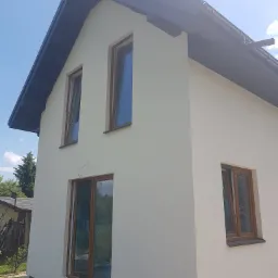 Nowo otynkowany dwukondygnacyjny dom z brązowymi ramami okiennymi i grafitowym dachem widziany z zewnątrz w słoneczny dzień.