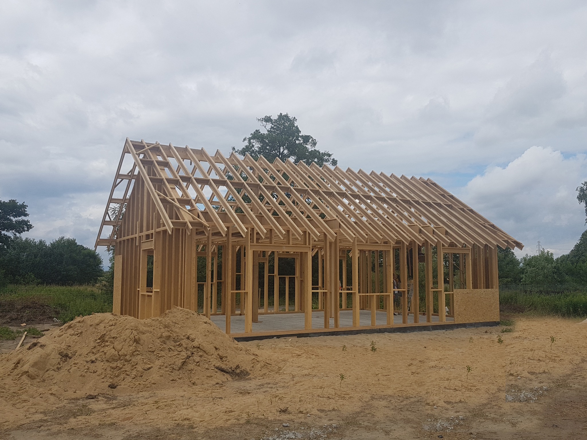 Wznoszenie konstrukcji drewnianej domu jednorodzinnego w stanie surowym otwartym, widoczny szkielet ścian i dachu, z hałdą piasku na pierwszym planie, na tle pochmurnego nieba.