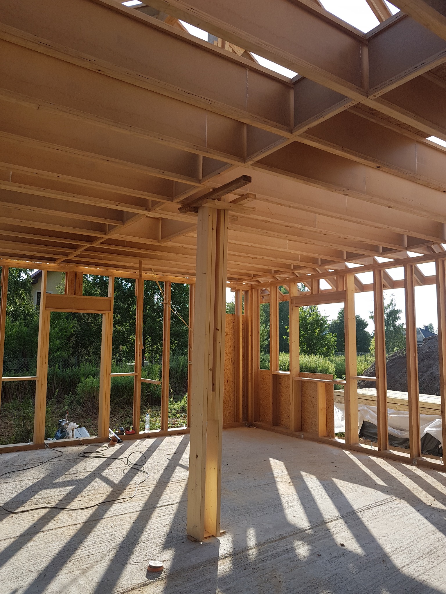 Wnętrze domu w trakcie budowy, widoczna drewniana konstrukcja szkieletowa ścian i stropu, z widocznymi narzędziami i materiałami budowlanymi, z zewnątrz widok na zieleń.