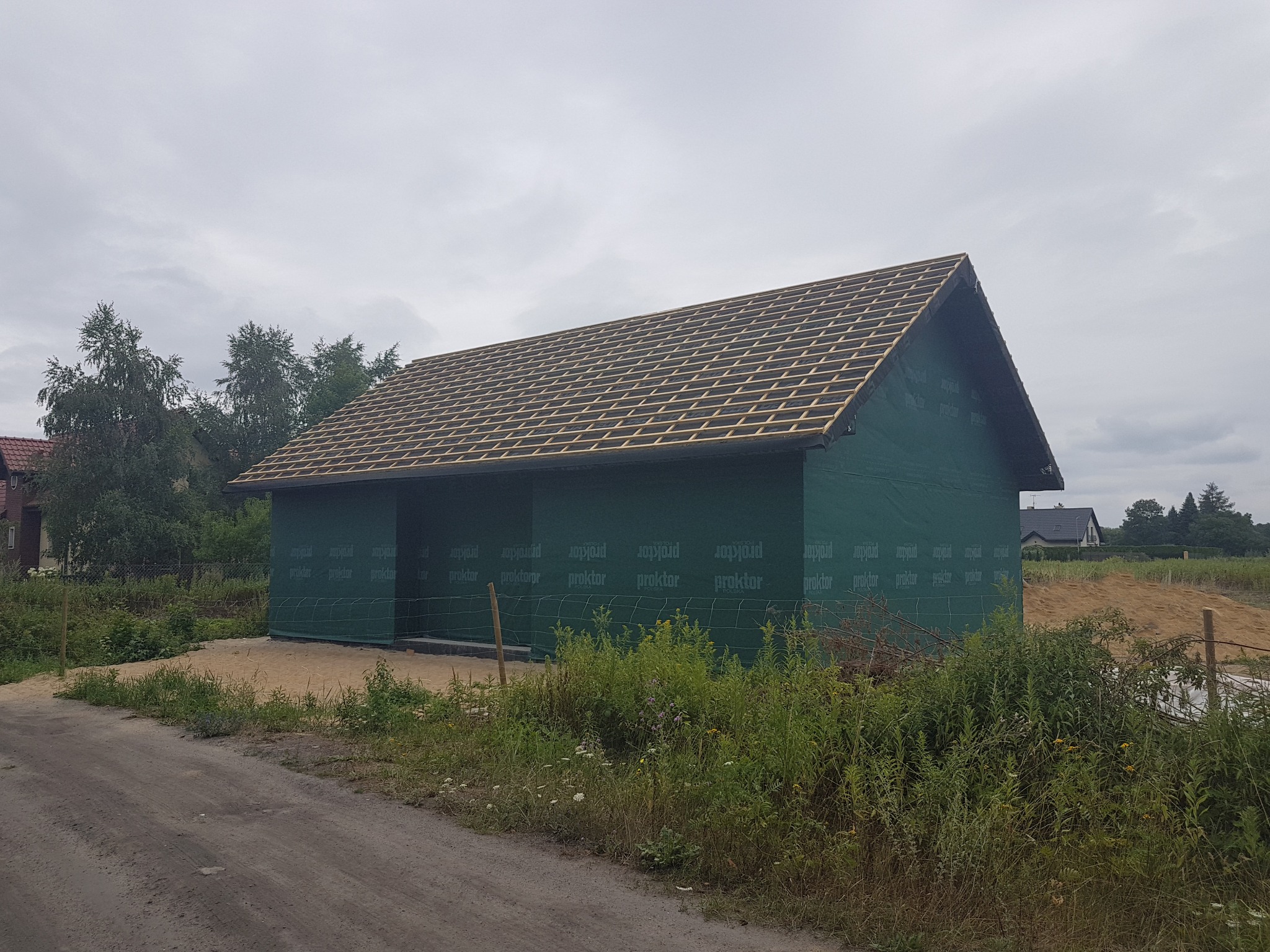 Surowy stan budowy domu jednorodzinnego z widoczną konstrukcją dachu i zieloną membraną ochronną na ścianach, otoczonego piaskiem i roślinnością.