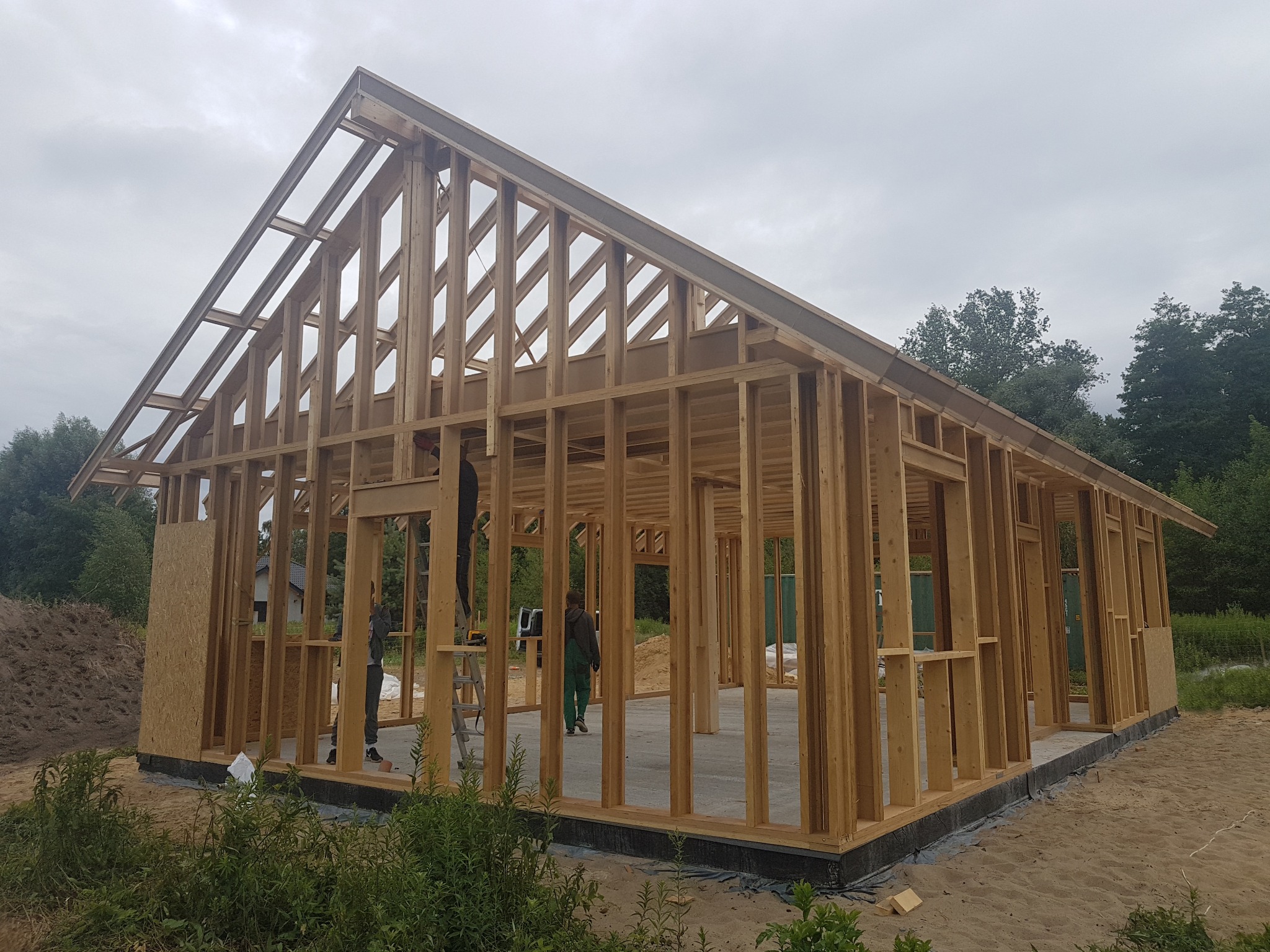 Konstrukcja szkieletowa domu w trakcie budowy, widoczne drewniane ramy ścian i dachu, betonowa podstawa i pracownicy w roboczych ubraniach.