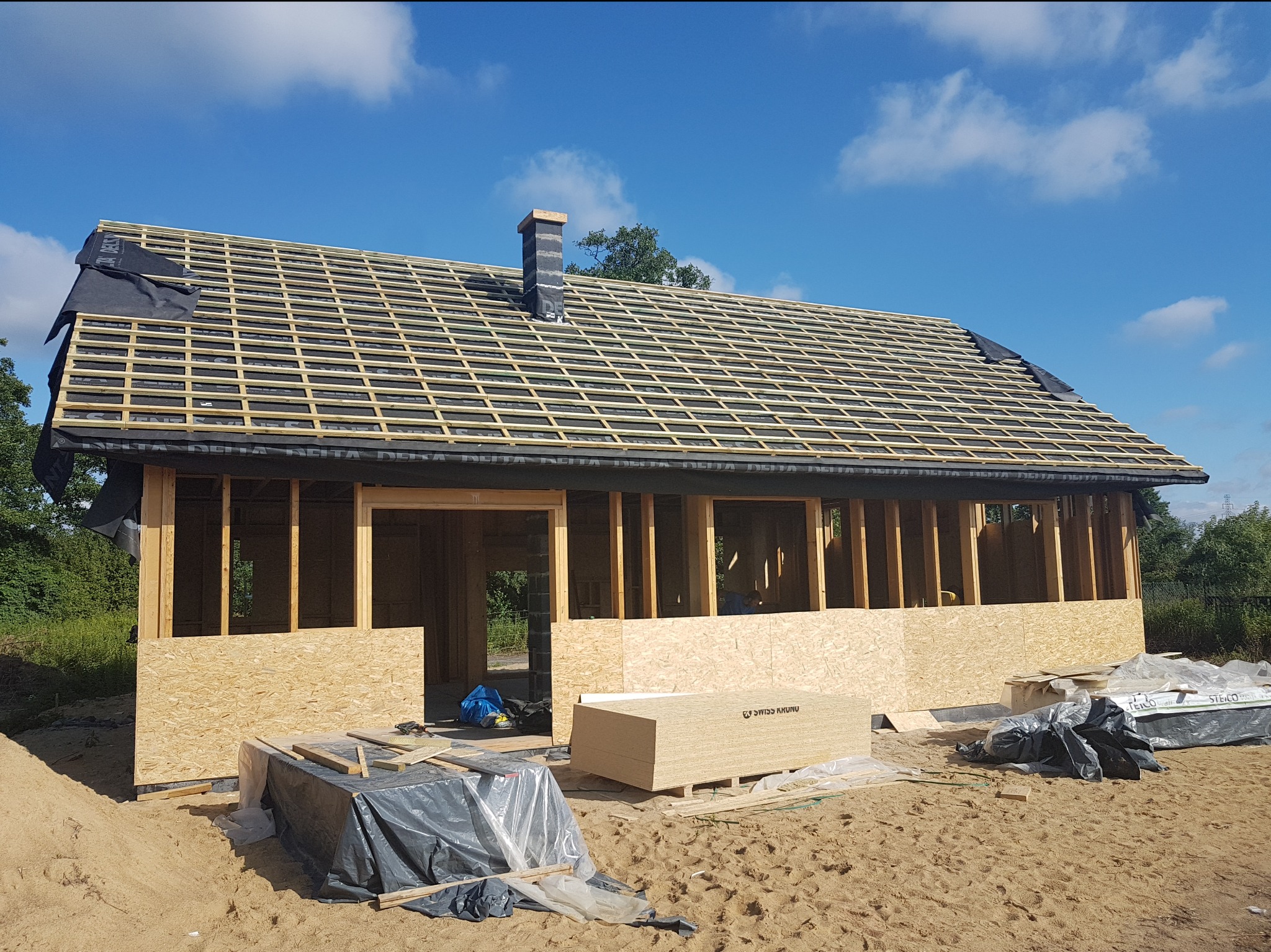 Wznoszenie konstrukcji domu z drewnianym szkieletem, widoczne płyty OSB, więźba dachowa z membraną, komin w trakcie budowy, materiały budowlane na piaszczystym podłożu.
