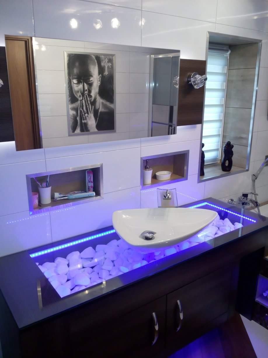 Nowoczesna łazienka z umywalką nablatową na szafce z białymi kamieniami podświetlonymi niebieskim światłem LED, lustro z wbudowanymi półkami i obrazem.