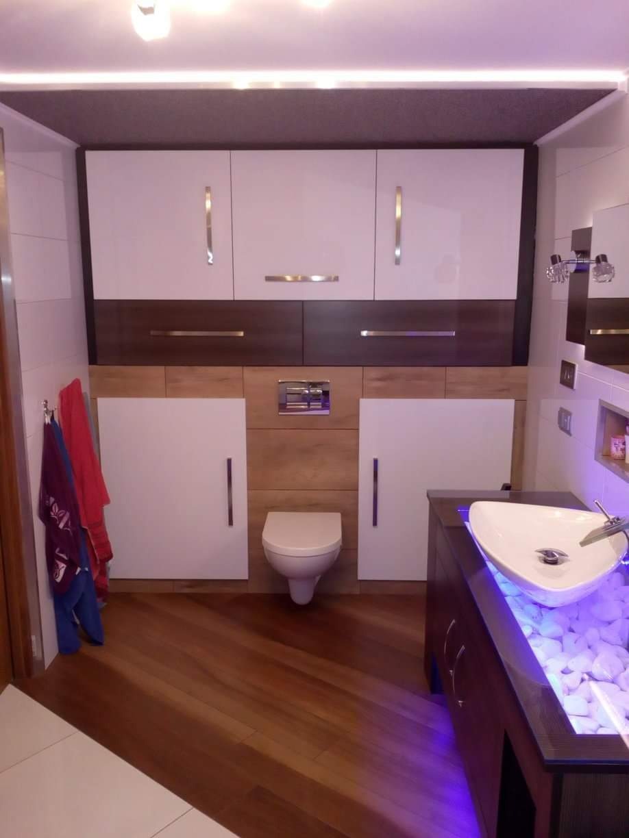 Nowoczesna łazienka z podwieszaną toaletą, szafkami w kolorze bieli i drewna, oraz umywalką podświetloną na fioletowo, otoczoną białymi kamieniami.