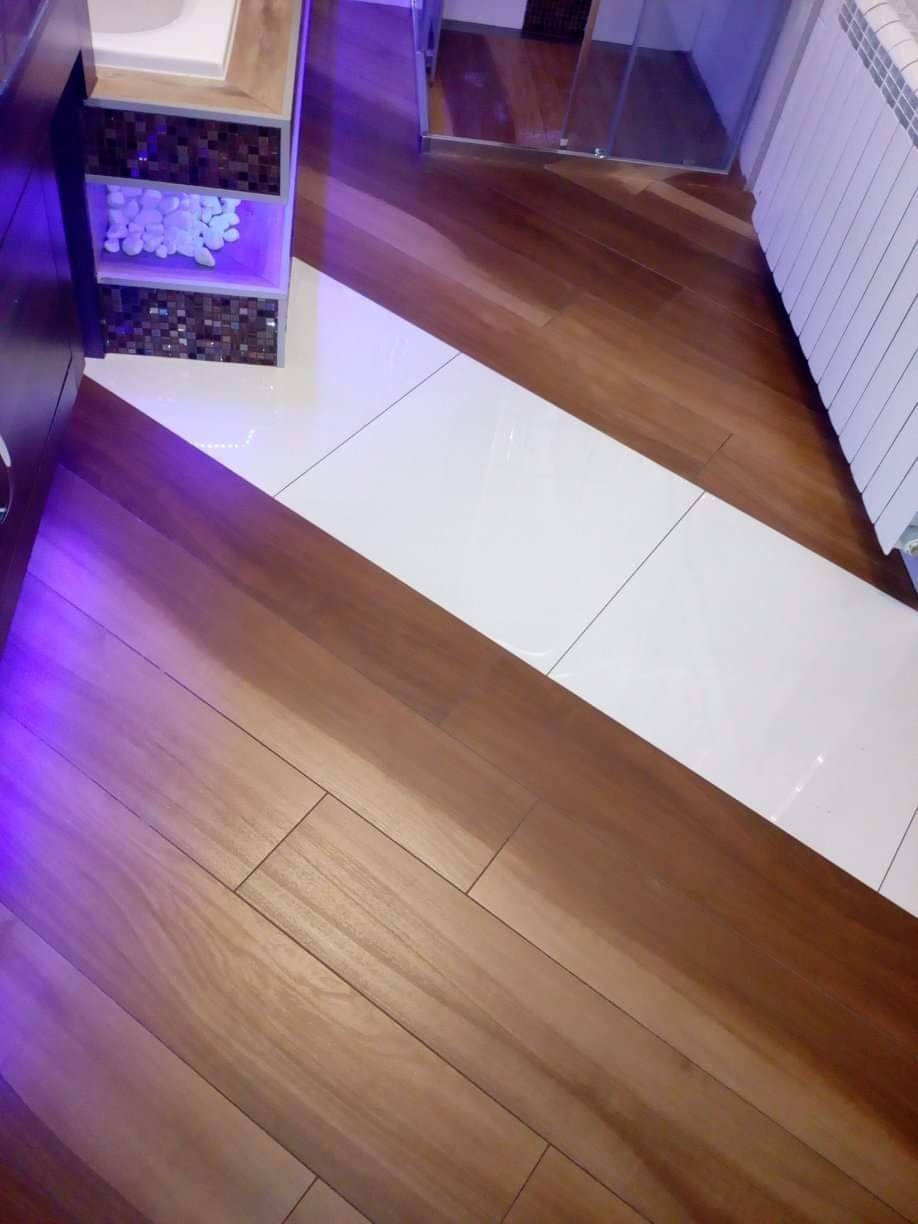 Ukośne ułożenie płytek podłogowych w łazience: drewnopodobne i białe, z szafką pod umywalką z mozaiką i dekoracyjnymi kamieniami oraz grzejnikiem żeberkowym w tle.