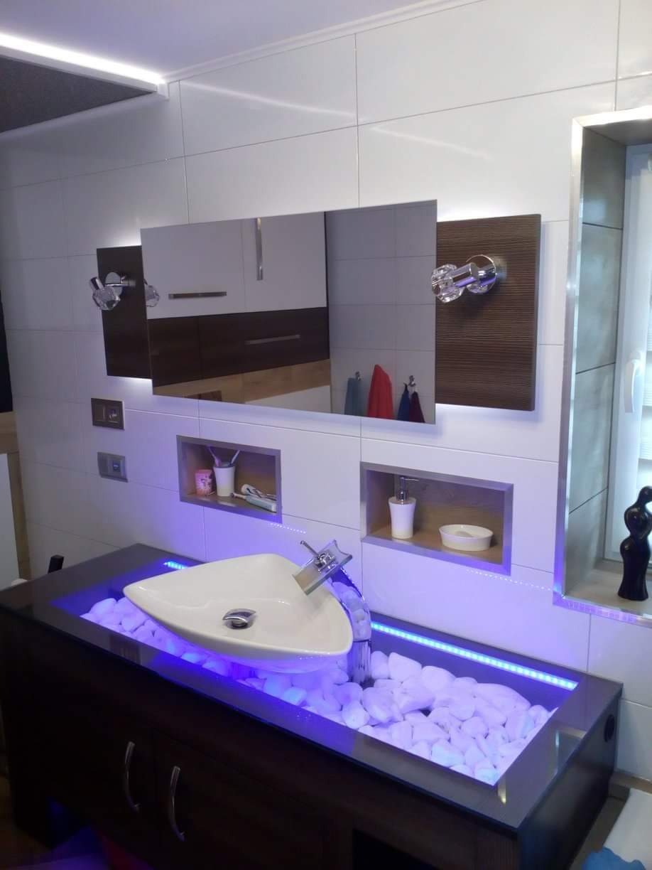 Nowoczesna łazienka z podświetlaną na niebiesko umywalką w kształcie trójkąta, otoczoną białymi kamieniami, wbudowana w szafkę z ciemnego drewna, z lustrem z kryształowymi uchwytami na ścianie...