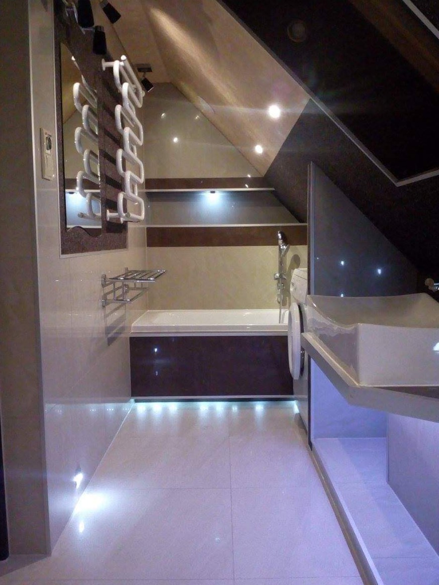 Nowoczesna łazienka na poddaszu z podświetlaną podłogą, wanną z ciemnym panelem i umywalką z szafką z subtelnym, fioletowym podświetleniem.