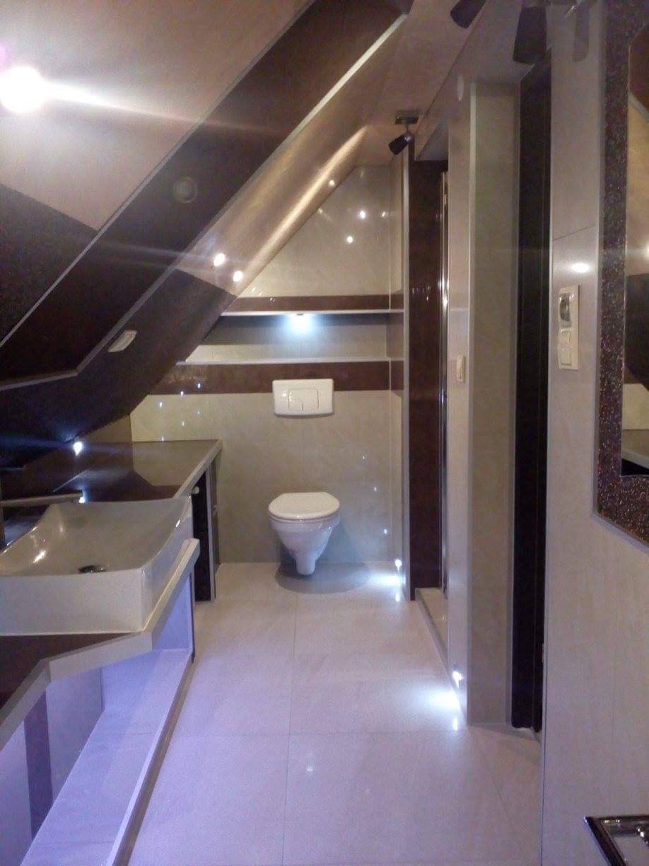 Nowoczesna łazienka na poddaszu z białymi płytkami, podświetlanym cokołem, umywalką nablatową i toaletą wiszącą.