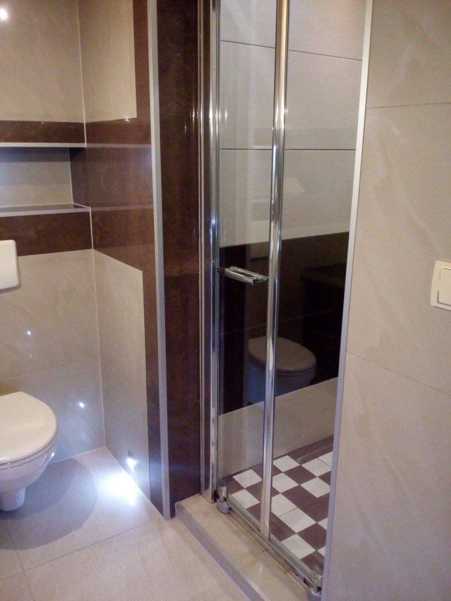 Nowoczesna łazienka z kabiną prysznicową z przesuwnymi drzwiami, brązowo-beżowe płytki na ścianach i podłodze w szachownicę, widoczne oświetlenie punktowe pod szafką.