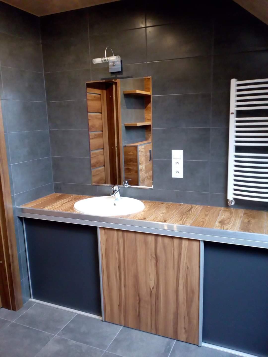 Nowoczesna łazienka z szarymi płytkami, drewnianym blatem i szafkami z przesuwnymi drzwiami, widoczny grzejnik drabinkowy i lustro z odbiciem drewnianych półek.