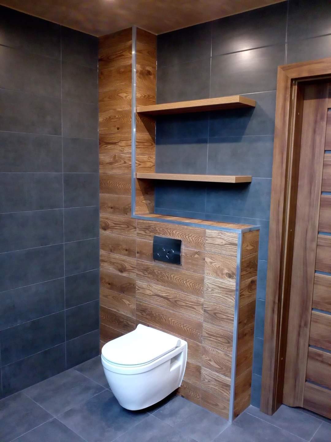 Nowoczesna łazienka z wiszącą toaletą, drewnianą zabudową i półkami, szare płytki na ścianach i podłodze, drewniane drzwi.
