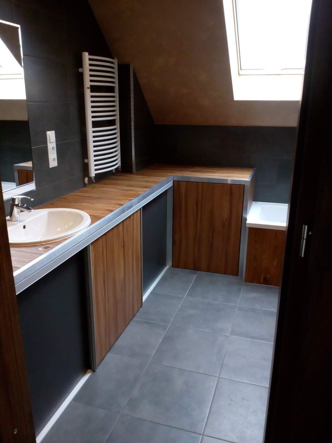 Nowoczesna łazienka na poddaszu z drewnianym blatem, szarą glazurą i grzejnikiem drabinkowym.