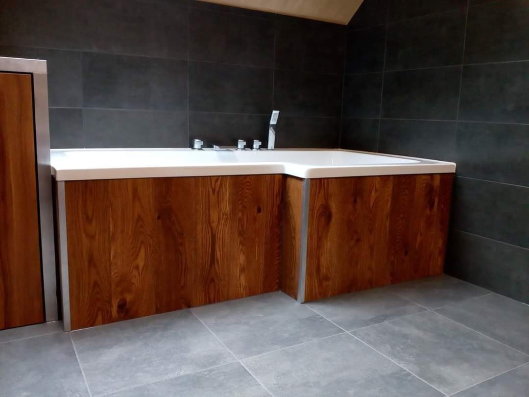 Biała narożna wanna obudowana drewnem w nowoczesnej łazience z ciemnymi płytkami na ścianach i szarymi płytkami na podłodze.